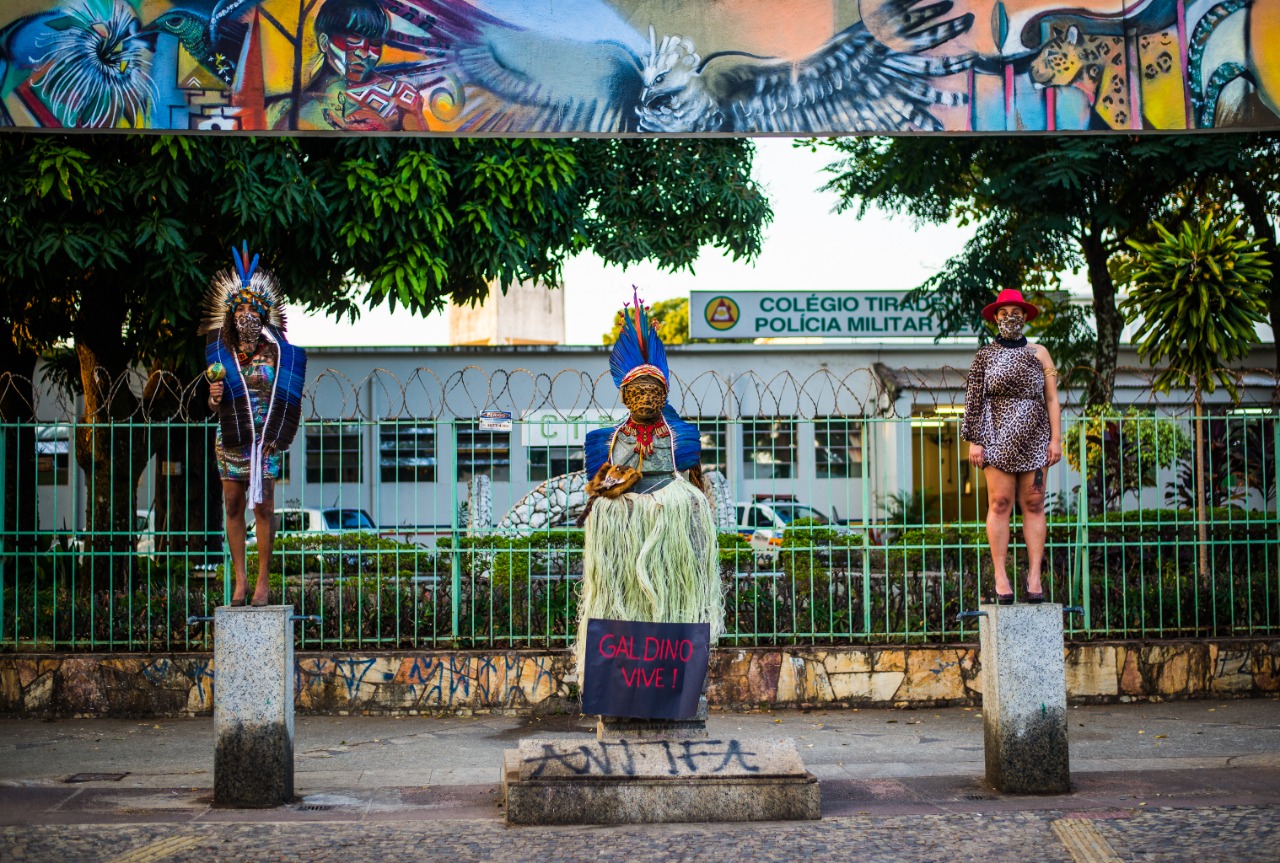 Duas mulheres de pé se colocam uma a cada lado de um busto de pedra vestido com roupas e acessórios indígenas. Elas estão equidistantes da estátua, olhando para a frente.