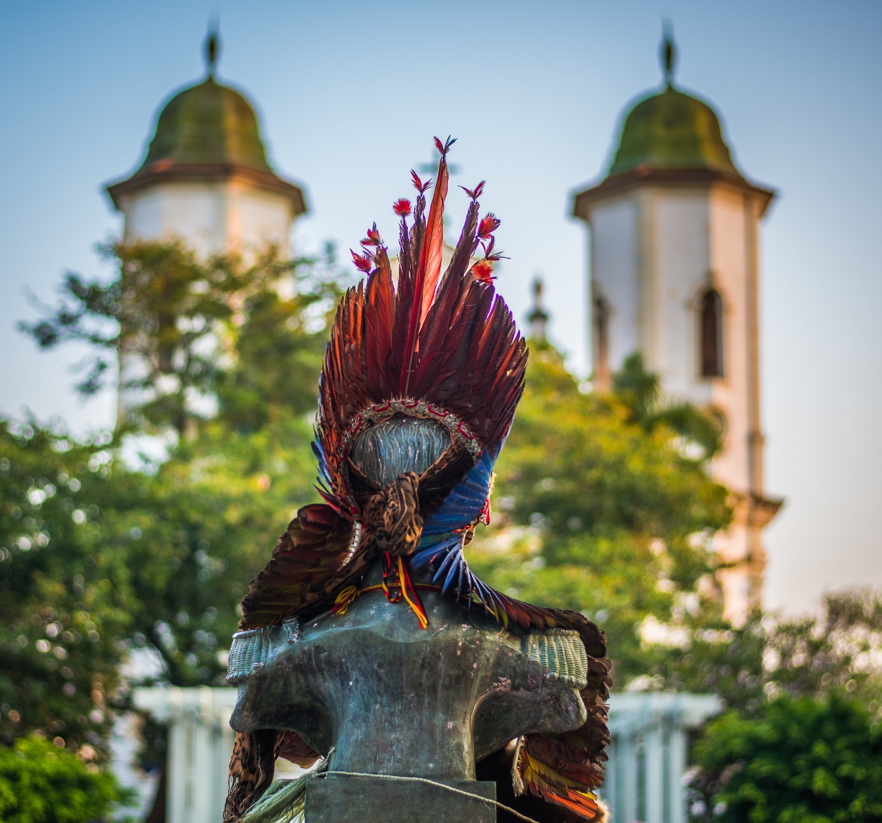 Imagem mostra busto de pedra de costas. Estátua tem um cocar vermelho.