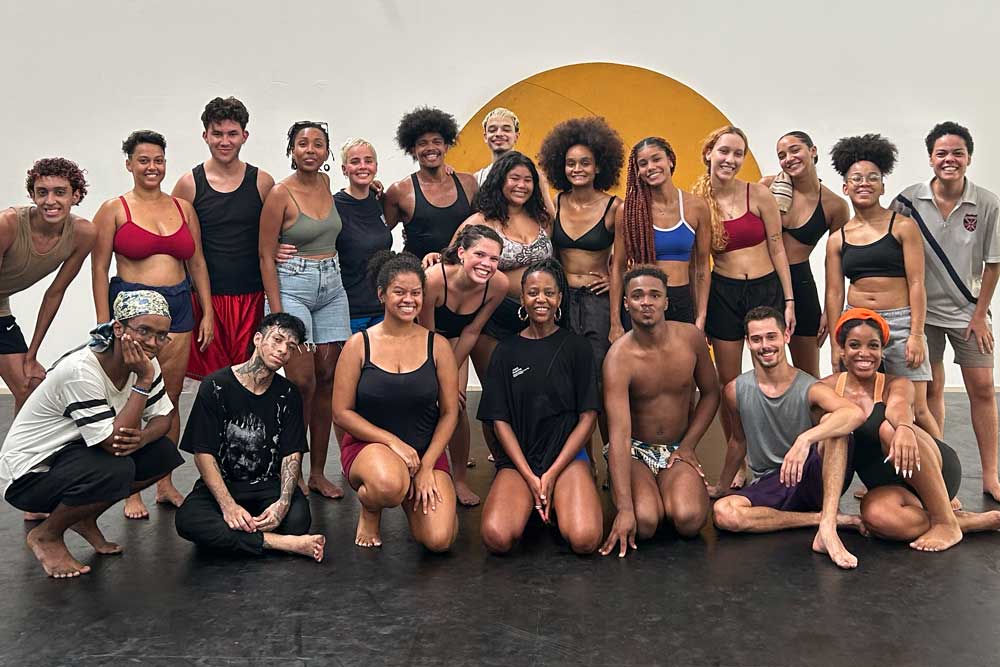 Grupo de aproximadamente 20 pessoas, diversas em idade, gênero e tons de pele, posando juntas em um estúdio de dança. Muitas estão sorrindo e vestem roupas confortáveis para prática corporal, como tops, shorts e camisetas. Algumas estão agachadas ou sentadas no chão, enquanto outras estão de pé. Ao fundo, uma parede branca com um círculo amarelo pintado no centro.